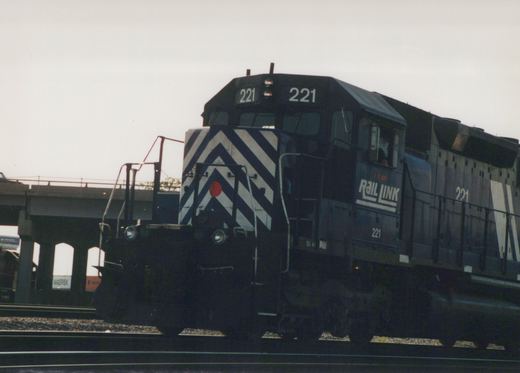 A black and white-striped diesel locomotive numbered 221 with I&M Rail Link lettering is moving on tracks under a concrete bridge.