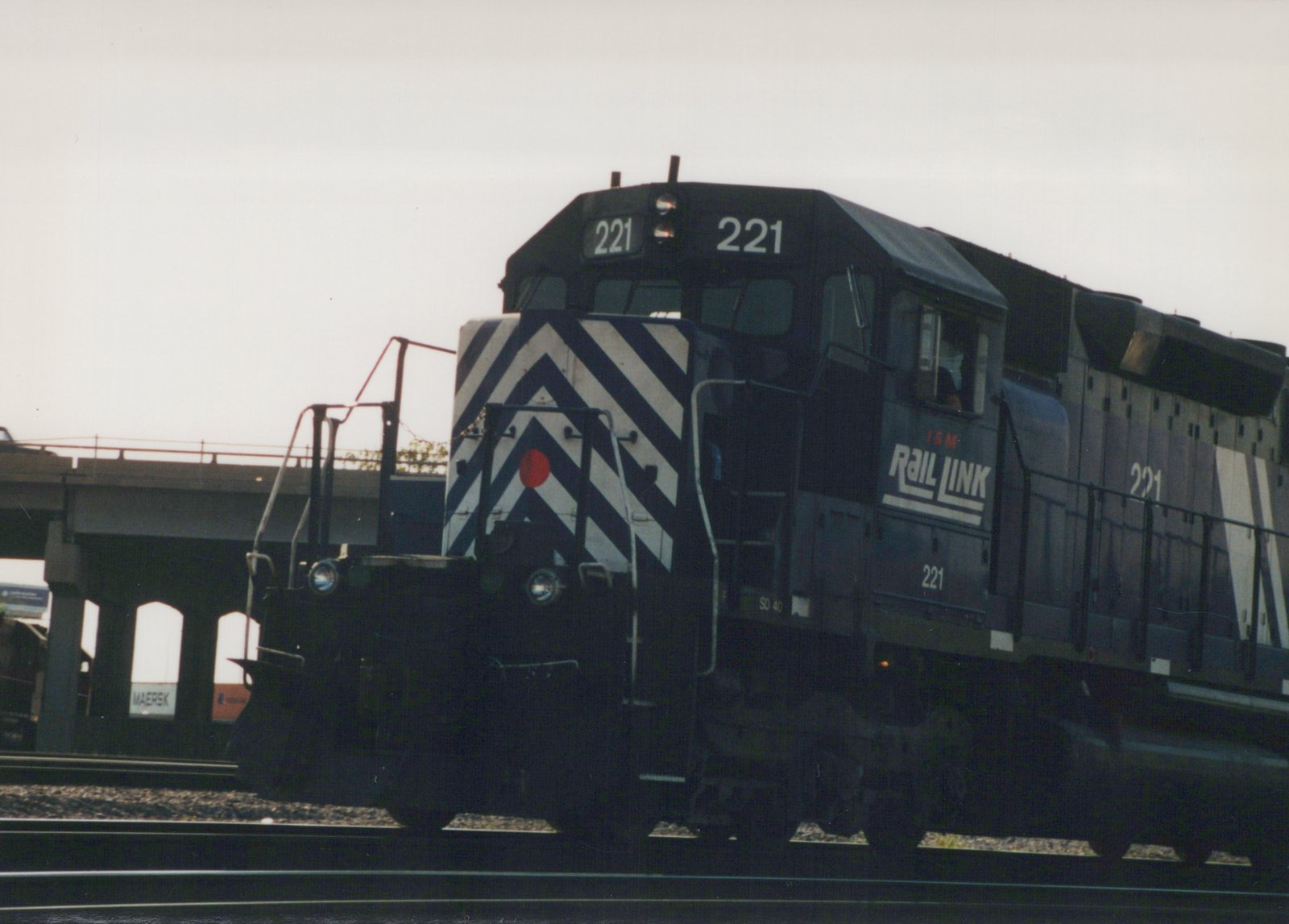 A black and white-striped diesel locomotive numbered 221 with I&M Rail Link lettering is moving on tracks under a concrete bridge.