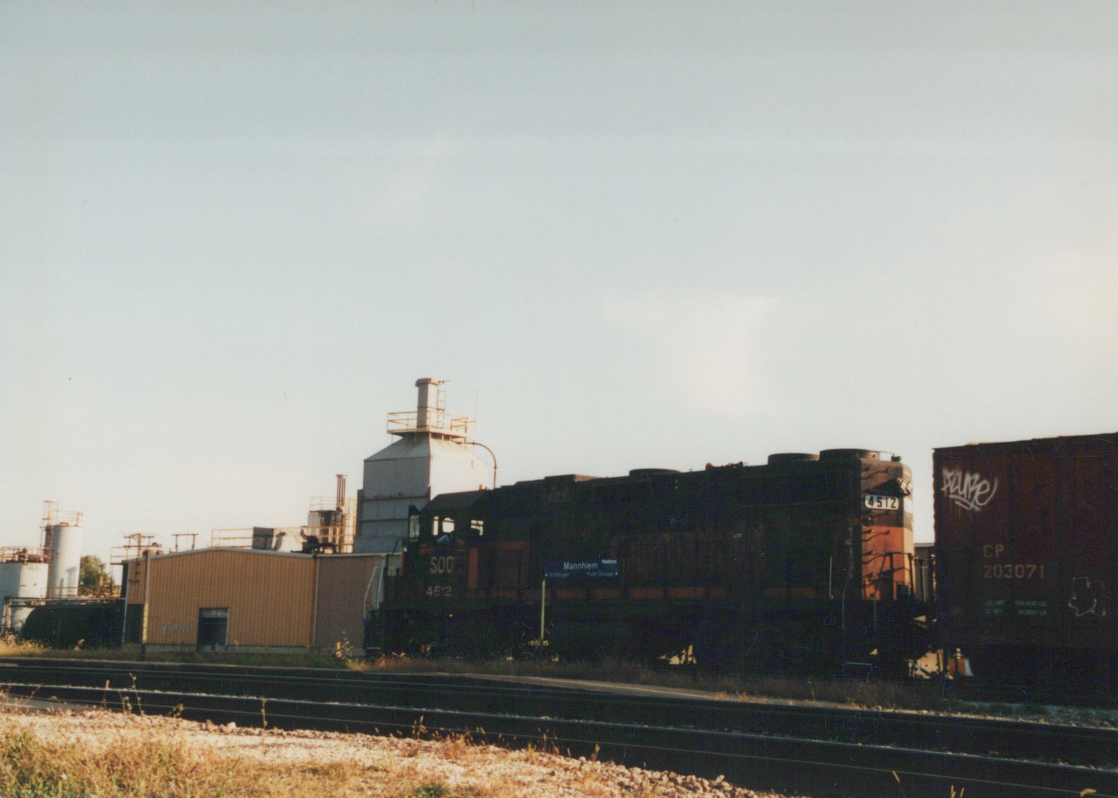 A dark green Soo Line locomotive with orange stripes, number 4512, is parked next to tracks with a blue 'Mannhiem' station sign and a CP freight car nearby.