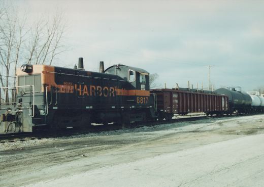 A black and orange diesel locomotive with the words Indian Harbor Belt and number 8817 on its side, pulling a line of freight cars in a yard under an overcast sky with bare trees.