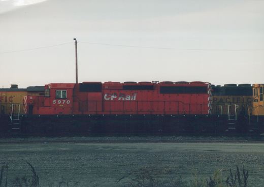 A red Canadian Pacific GP9 diesel locomotive numbered 5970 sits on tracks in a rail yard, with a yellow locomotive partially visible behind it.