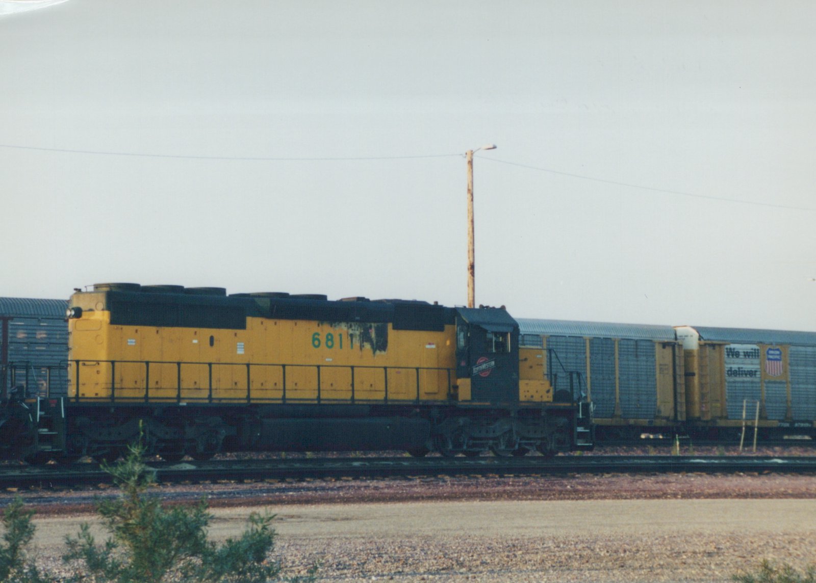 A yellow and black Chicago and North Western diesel locomotive numbered 6811 is shown on a railroad track with freight cars, under an overcast sky.