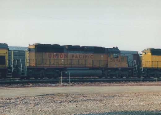 A side view of a Union Pacific yellow and black GP9 diesel locomotive, number 3694, parked in a railroad yard next to other cars and a second locomotive.