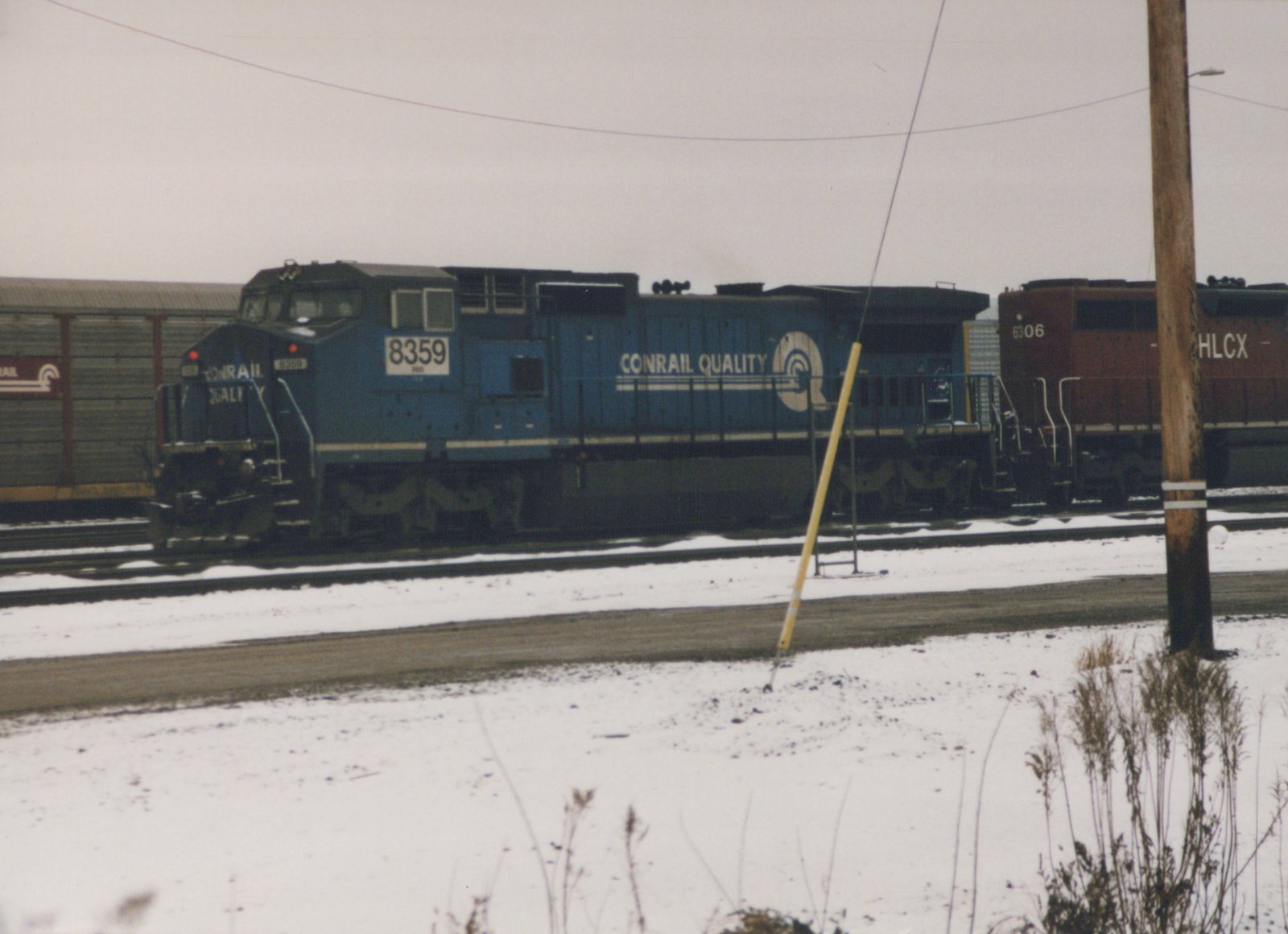 A side view of two diesel locomotives in a snow-covered yard, with the foreground train in blue paint labeled 'CONRAIL QUALITY' and numbered 8359, and a maroon train labeled 'HLCX 6306' behind it.