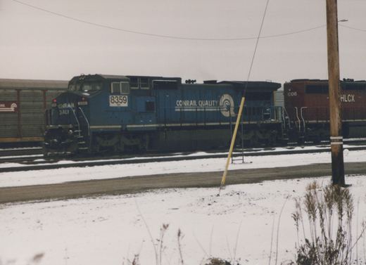 A side view of two diesel locomotives in a snow-covered yard, with the foreground train in blue paint labeled 'CONRAIL QUALITY' and numbered 8359, and a maroon train labeled 'HLCX 6306' behind it.