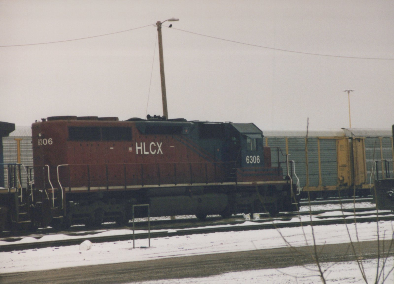 A maroon and blue diesel locomotive with HLCX lettering and number 6306 sits on the tracks in a snowy yard.