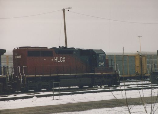 A maroon and blue diesel locomotive with HLCX lettering and number 6306 sits on the tracks in a snowy yard.