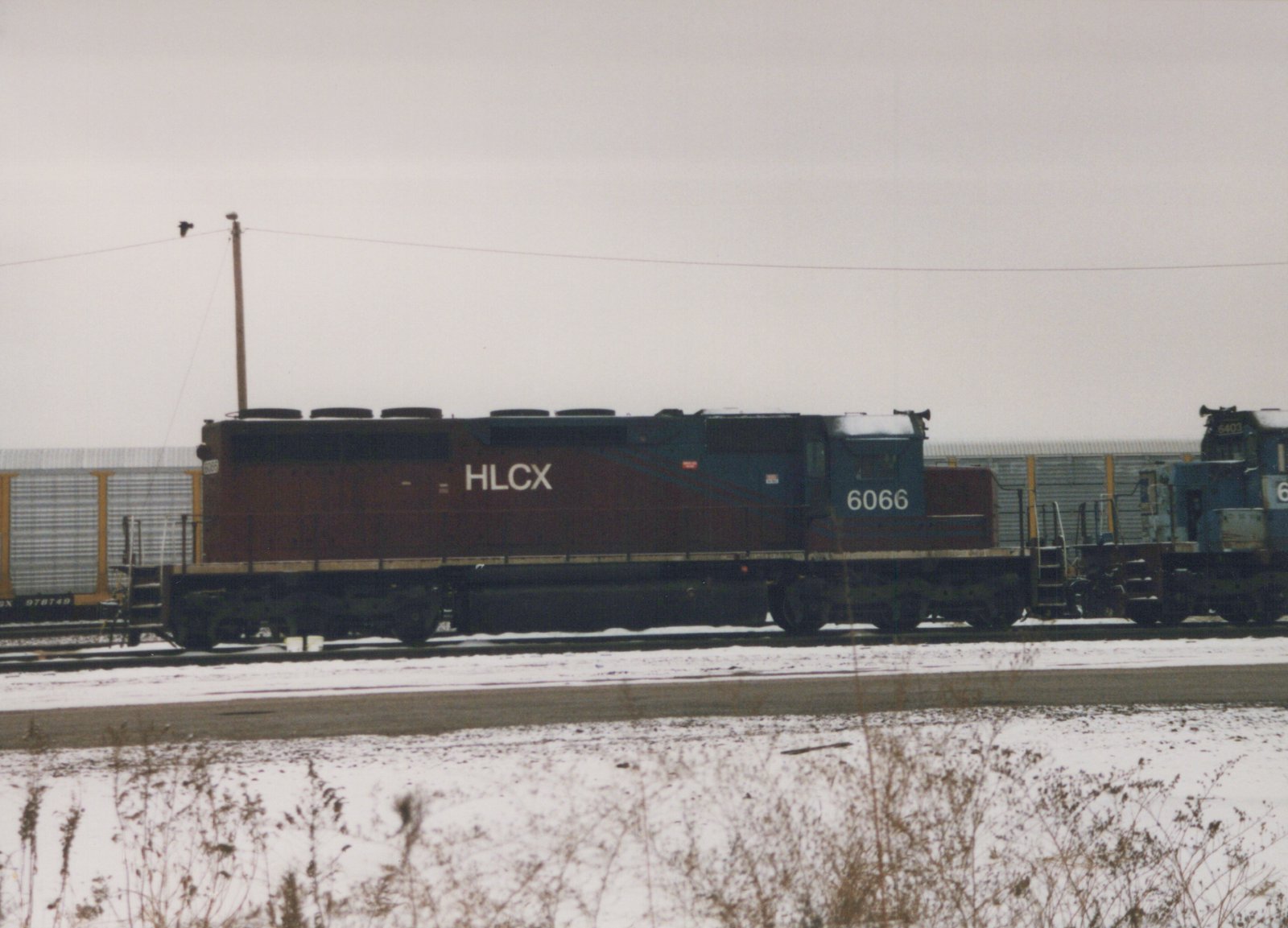 Two HLCX diesel locomotives with numbers 6066 and 6403 are parked side by side in a snowy rail yard, with a freight car marked 978749 nearby.