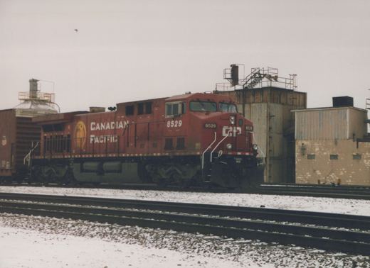 A red Canadian Pacific locomotive, number 8529, on tracks with snow on the ground and industrial buildings in the background.