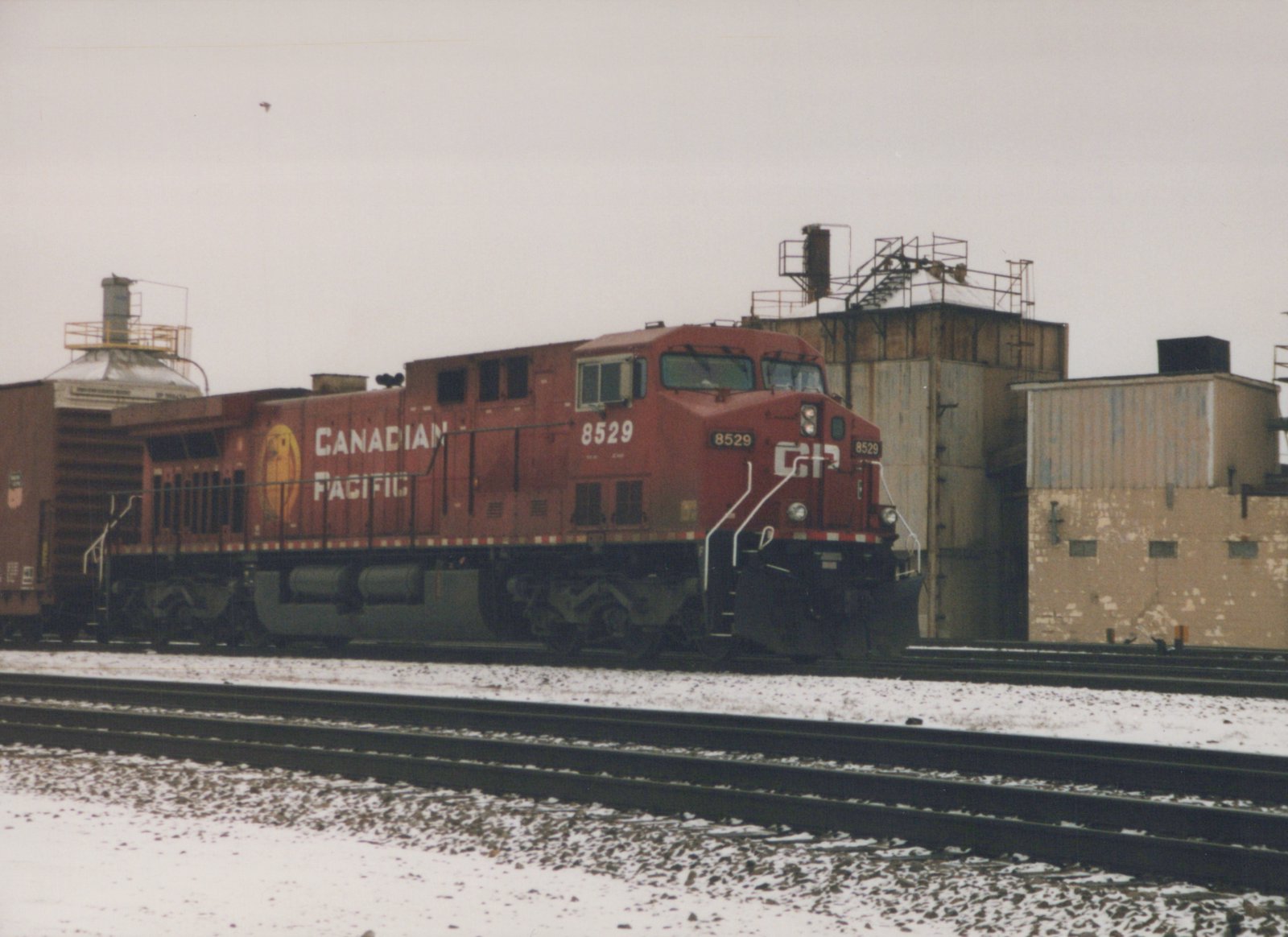 A red Canadian Pacific locomotive, number 8529, on tracks with snow on the ground and industrial buildings in the background.