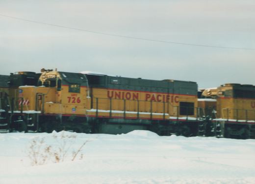 A yellow and gray Union Pacific diesel locomotive with the number 726 is parked in a snowy rail yard, with other locomotives visible in the background.
