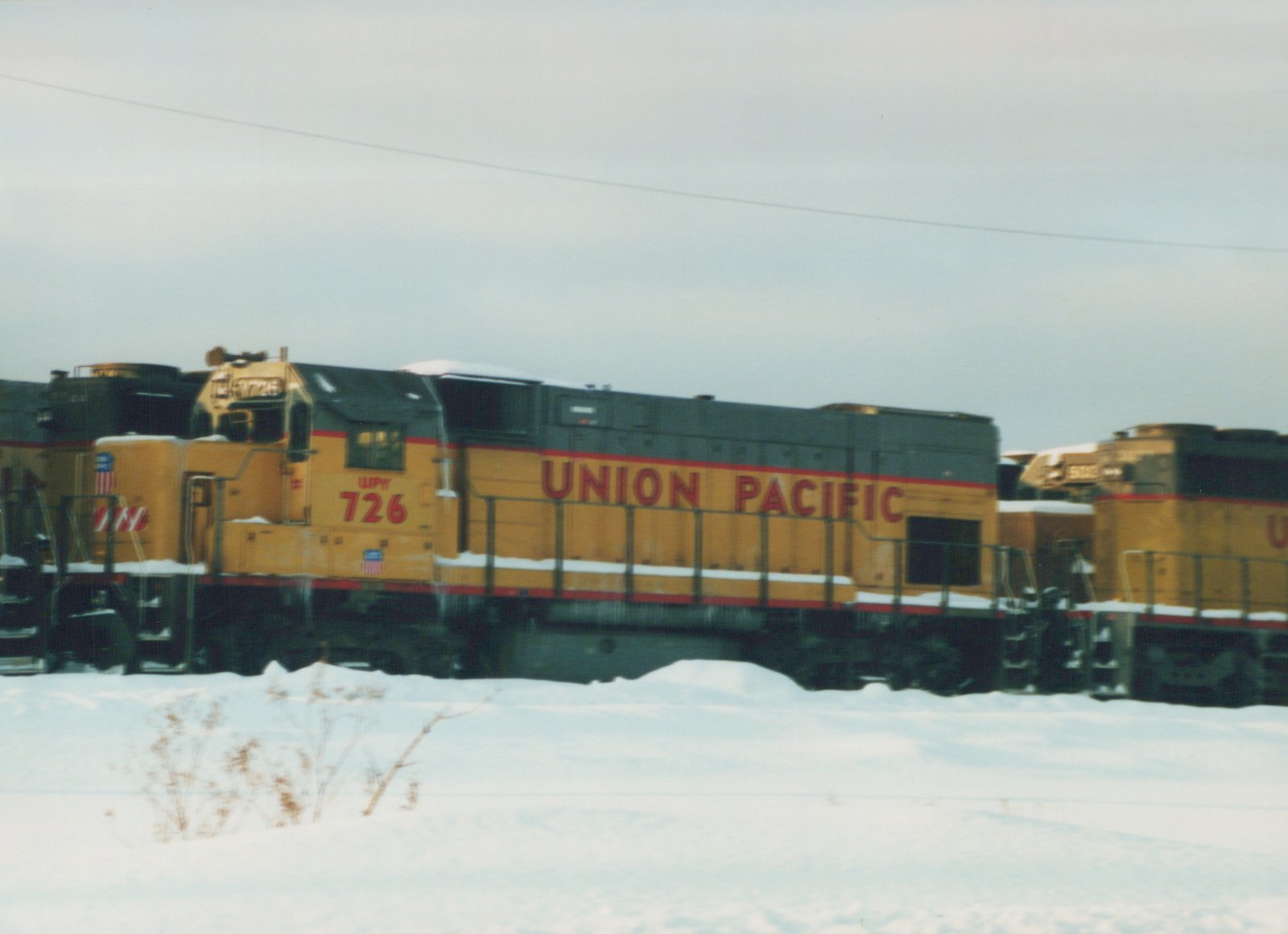 A yellow and gray Union Pacific diesel locomotive with the number 726 is parked in a snowy rail yard, with other locomotives visible in the background.