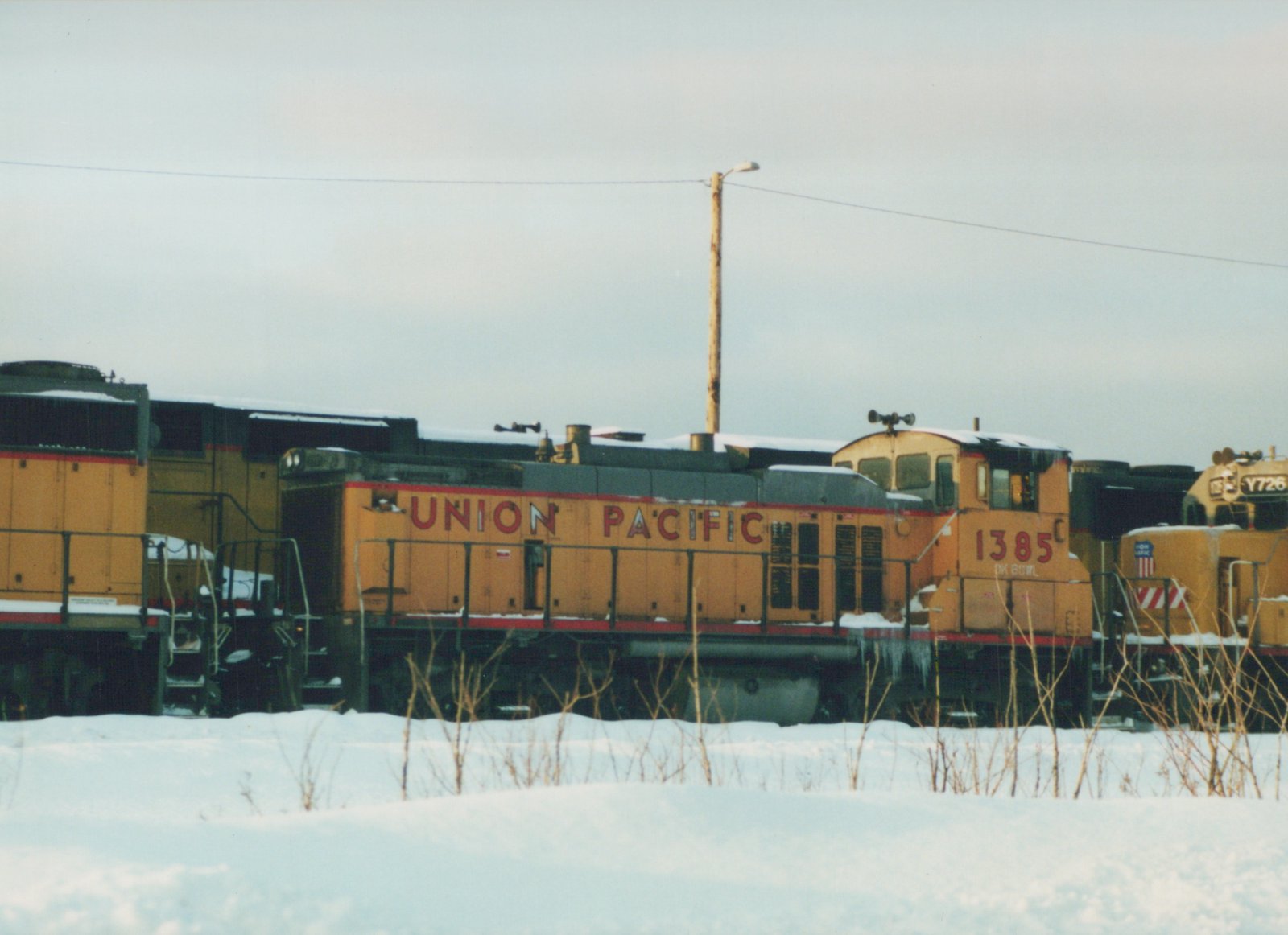 A side view of a yellow Union Pacific diesel locomotive with the number 1385 on its cab, parked in a snowy yard with another locomotive visible in the background.