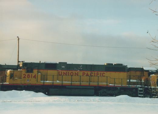 Side view of a yellow and black Union Pacific diesel locomotive, number 2814, parked on tracks next to a snow-covered field with bare tree branches visible in the corner.