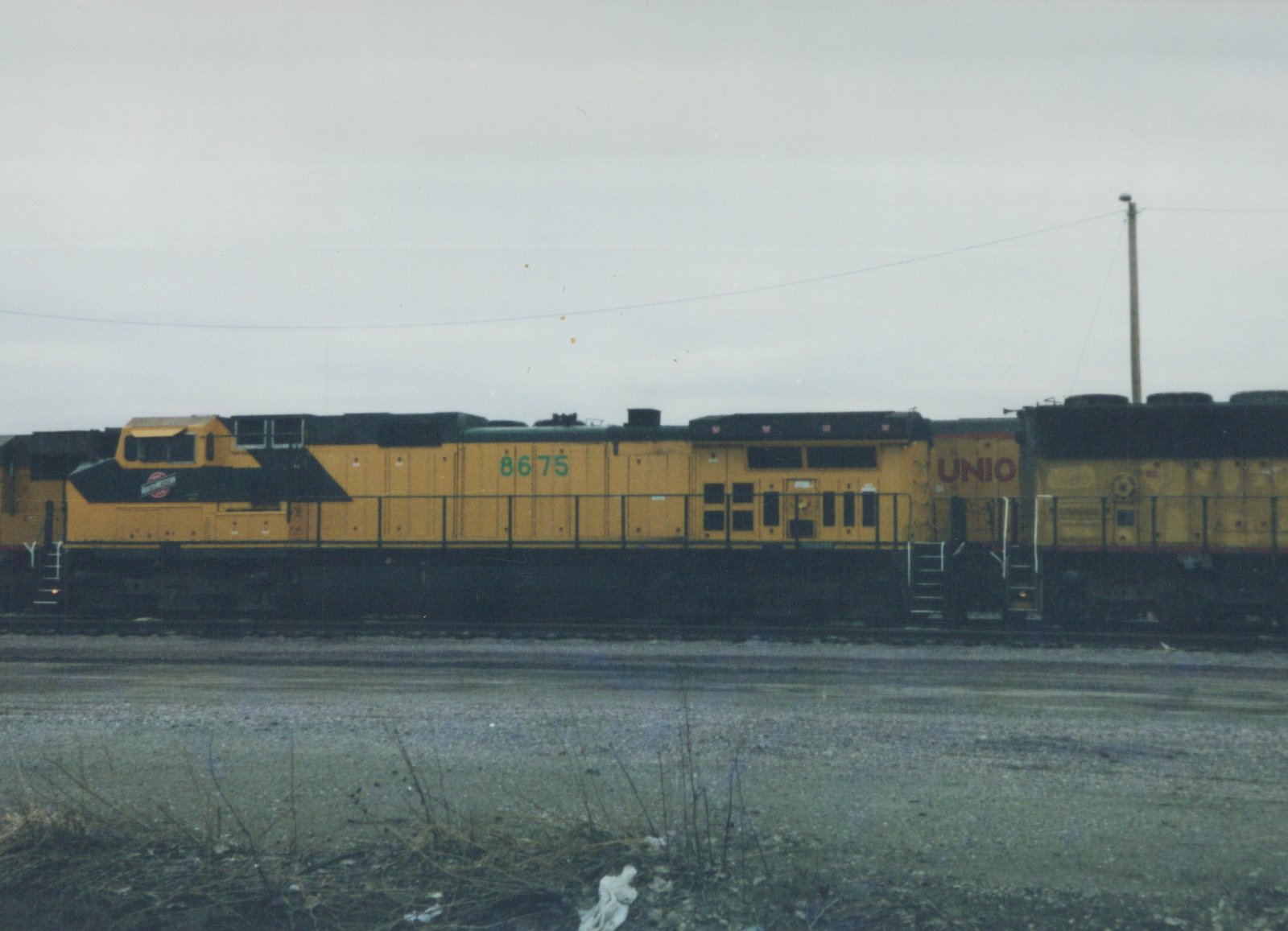 A yellow and black diesel locomotive with the number 8675 and partial lettering 'UNIO' is parked on a set of tracks in a gravel yard under an overcast sky.