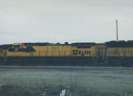 A yellow and black diesel locomotive with the number 8675 and partial lettering 'UNIO' is parked on a set of tracks in a gravel yard under an overcast sky.
