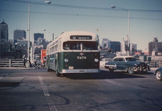 Front three-quarter view of a green and cream Chicago Transit Authority bus, number 7279, on a city street bridge with 1950s cars and skyscrapers behind it.