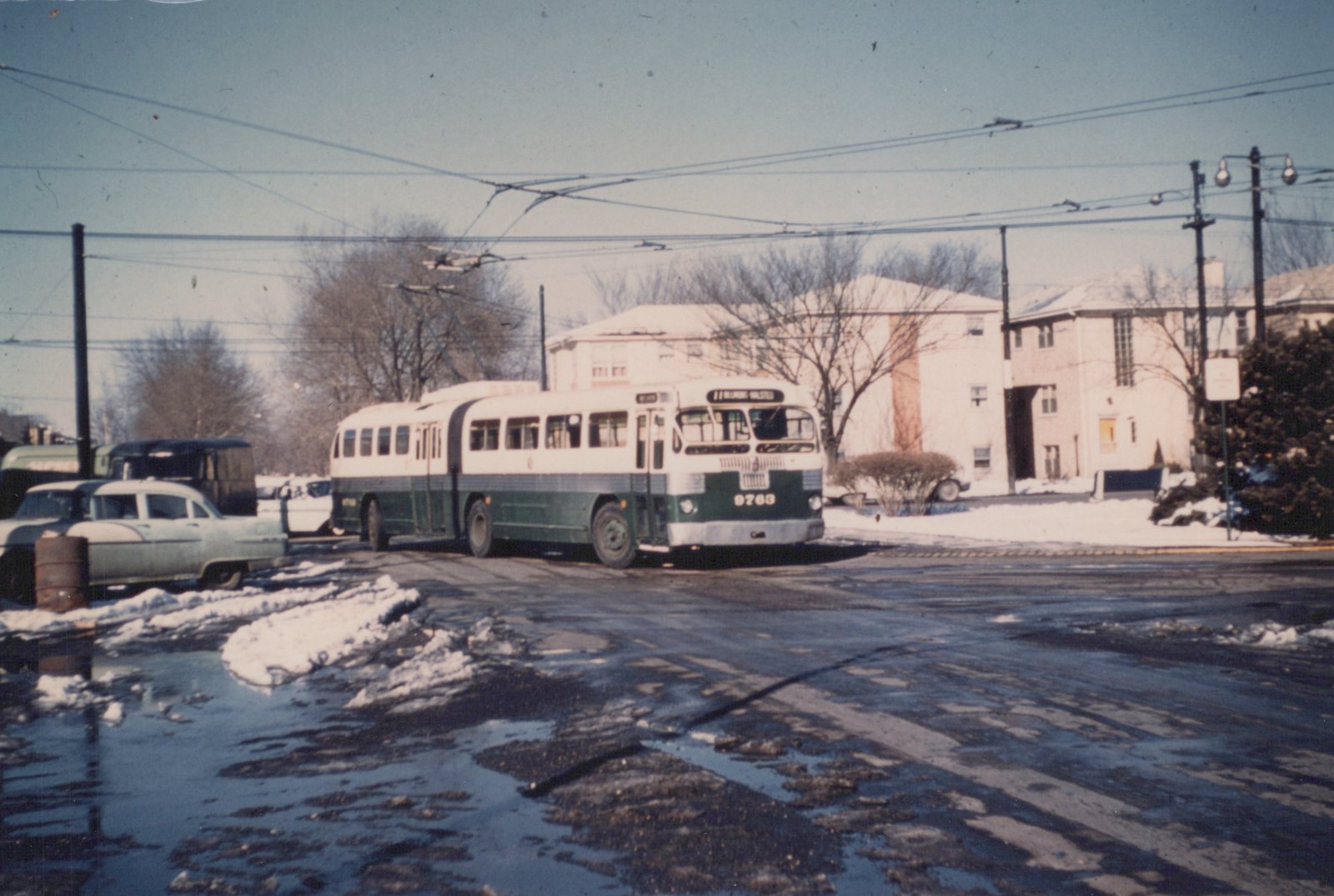 A green and cream CTA trolleybus, number 9763, is parked on a snowy street with overhead wires, near a 1950s car and apartment buildings.
