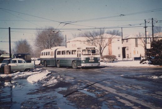 A green and cream CTA trolleybus, number 9763, is parked on a snowy street with overhead wires, near a 1950s car and apartment buildings.