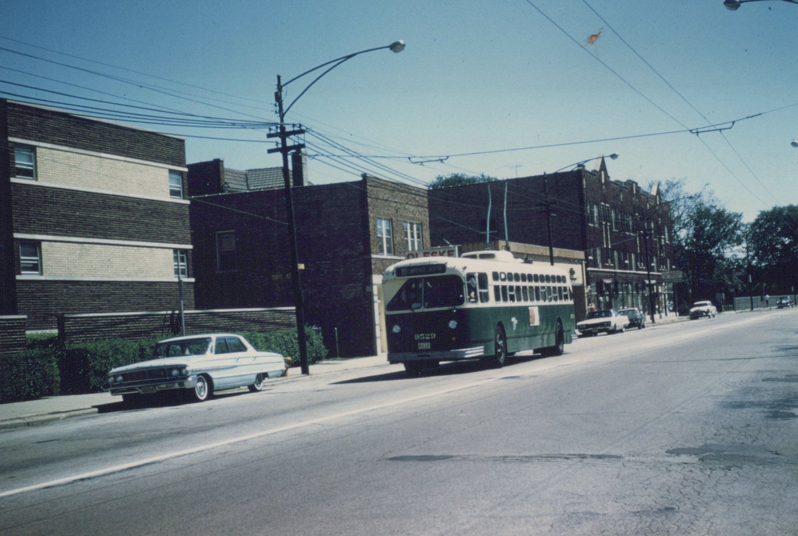 A green and cream trolleybus with the number 9529 and destination 'Lawrence' drives down a sunlit Chicago street lined with brick buildings and parked cars.