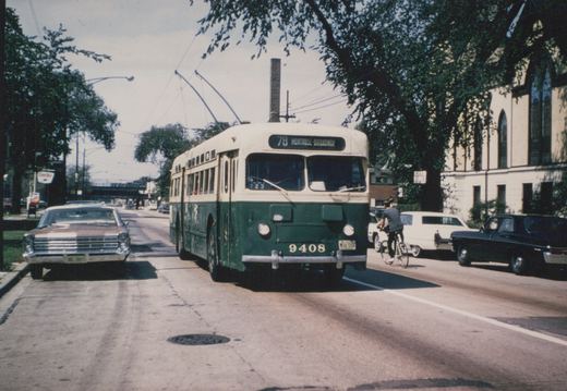 A green and cream trolleybus numbered 9408 is driving down a tree-lined city street in summer, with a cyclist and several cars nearby.