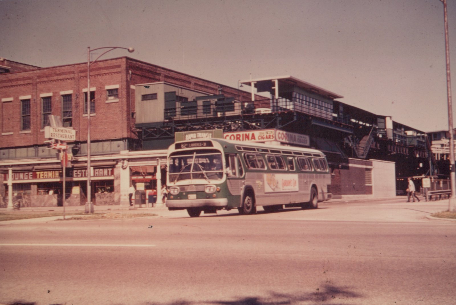 A green and cream Chicago Transit Authority bus drives down a city street under an elevated train station, with a brick building and restaurant sign visible in the background.