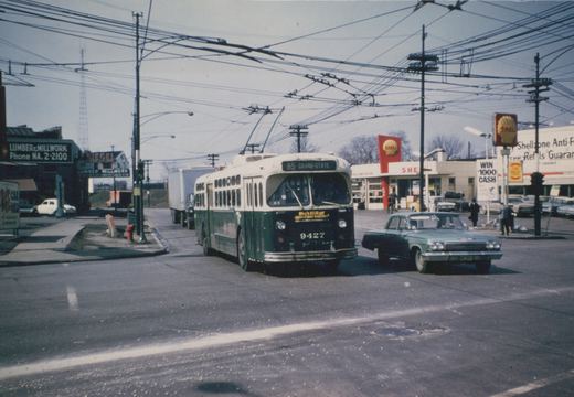 A green and cream trolleybus with the number 9427 is turning at an intersection with a Shell gas station in the background, under a network of overhead wires, with a green car and pedestrians nearby.