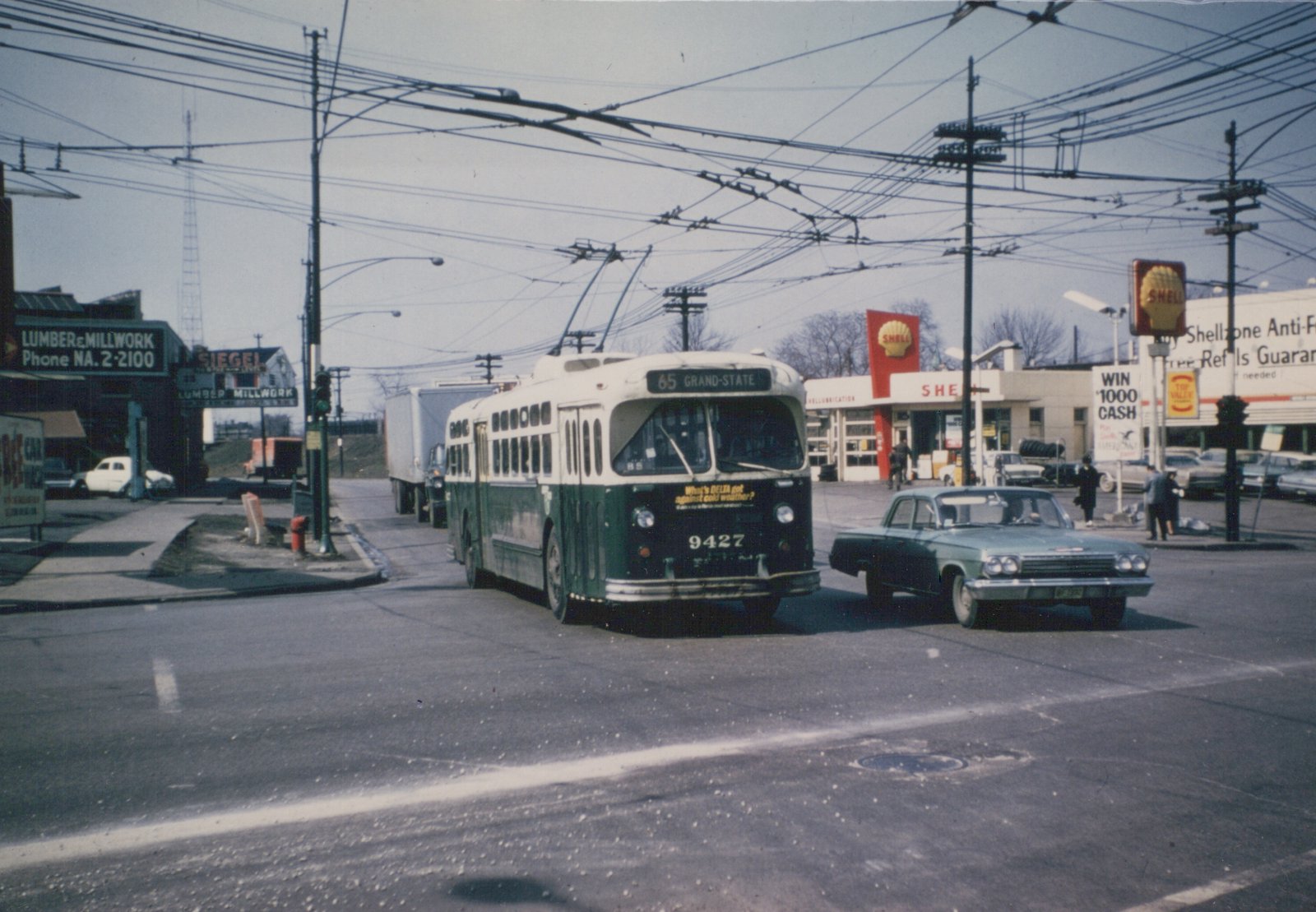 A green and cream trolleybus with the number 9427 is turning at an intersection with a Shell gas station in the background, under a network of overhead wires, with a green car and pedestrians nearby.