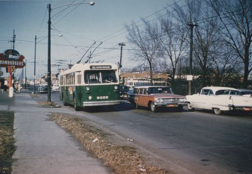 A green and cream trolleybus with the number 9355 and route 85 Central-Harrison is stopped on a street, with several vintage cars around it and bare trees in the background.
