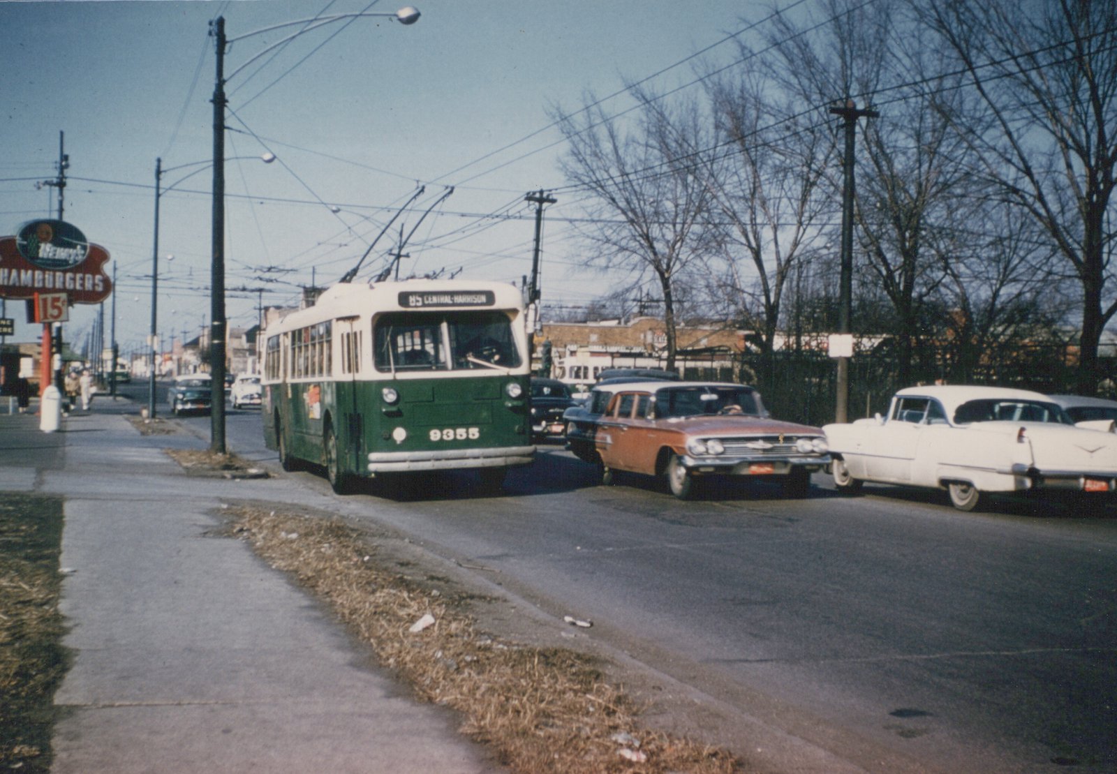 A green and cream trolleybus with the number 9355 and route 85 Central-Harrison is stopped on a street, with several vintage cars around it and bare trees in the background.