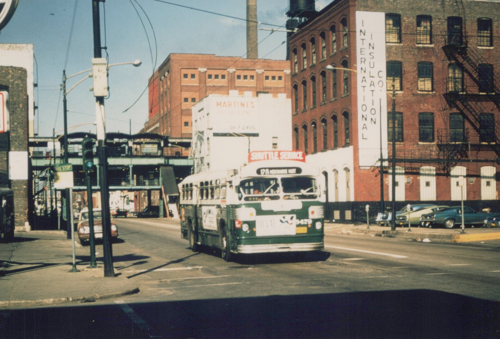 A green and cream CTA bus numbered 3429 labeled 'SHUTTLE SERVICE' is turning at an intersection with elevated tracks and brick buildings in the background.