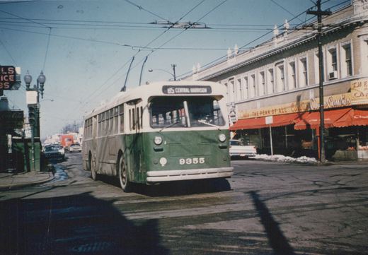Front three-quarter view of a green and cream CTA trolleybus, number 9355, on route 85 Central-Harrison, with overhead wires and storefronts in the background.
