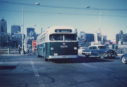 A vintage green and cream Chicago Transit Authority bus, number 7279, is shown on a city street with a 1950s car nearby and skyscrapers in the distance.