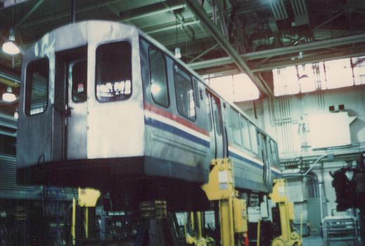 A silver rapid transit car with red and blue stripes is raised on yellow lifts inside a large industrial shop with high ceilings and windows.