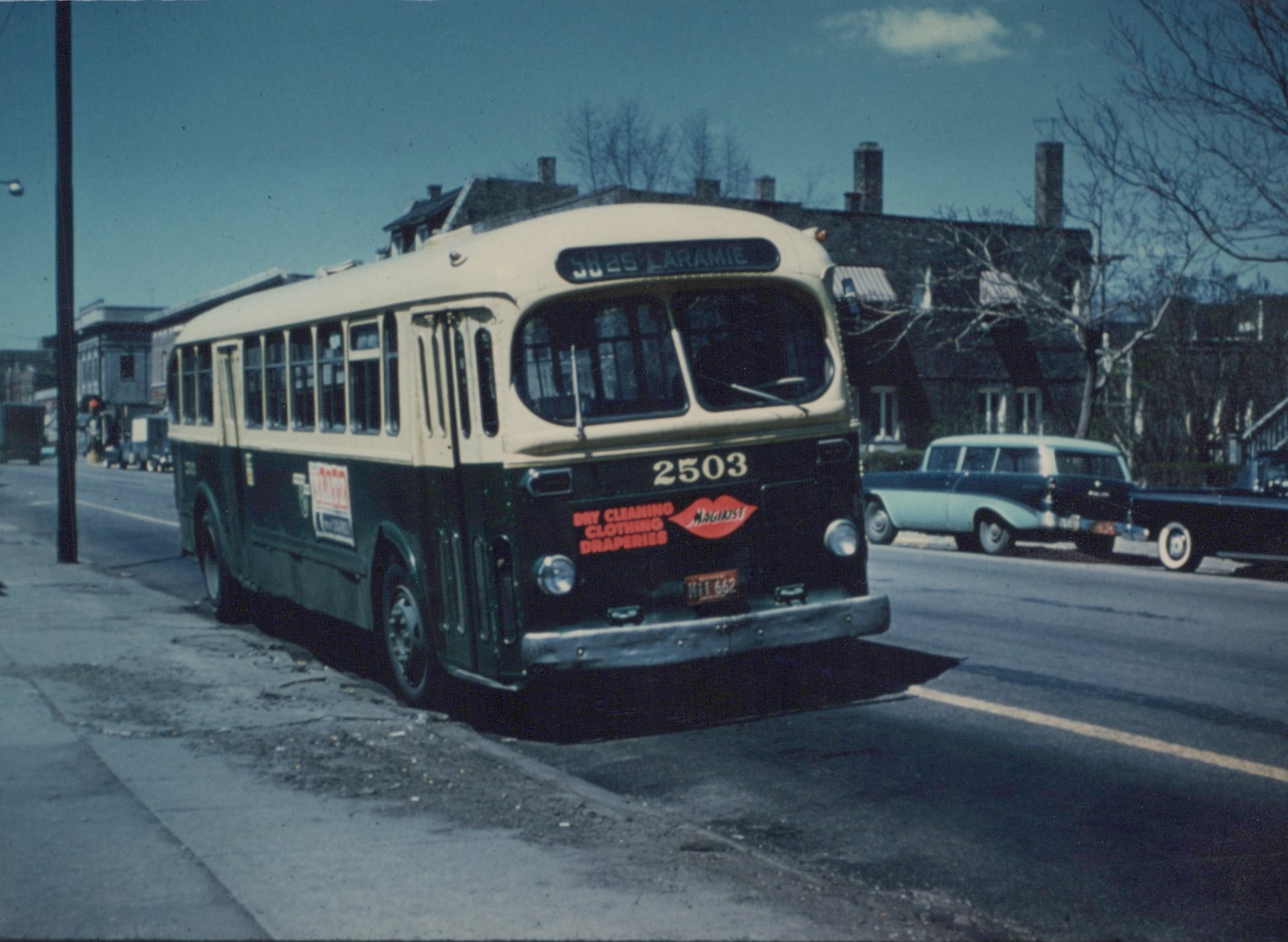 A vintage green and cream bus with the number 2503 on its front is parked on a city street, with a 1950s station wagon visible in the background.
