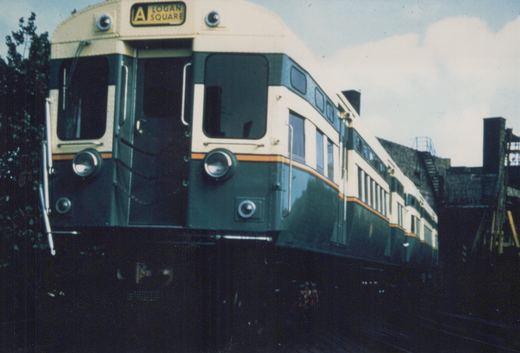 Front three-quarter view of a green and cream Chicago Transit Authority 2000-series rapid transit car with an 'A LOGAN SQUARE' destination sign, running on an elevated structure under an overcast sky.