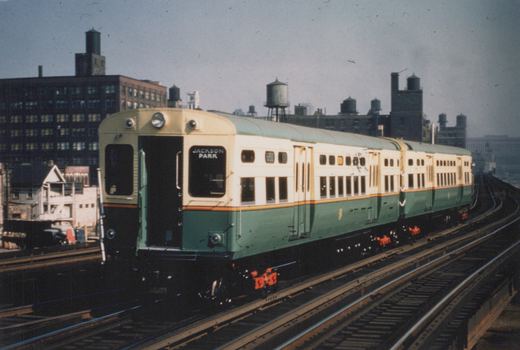 A green and cream Chicago Transit Authority rapid transit train, car number 6150, travels on an elevated track with a destination sign reading 'JACKSON PARK', set against an urban skyline with water towers.