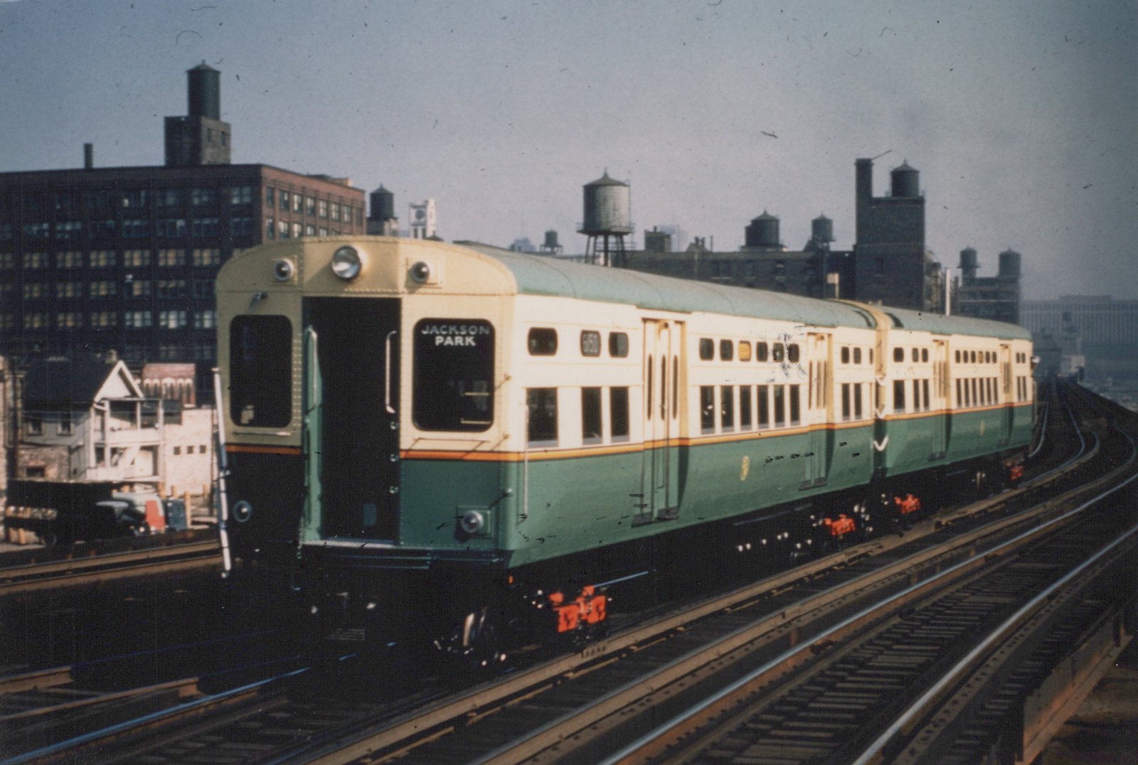 A green and cream Chicago Transit Authority rapid transit train, car number 6150, travels on an elevated track with a destination sign reading 'JACKSON PARK', set against an urban skyline with water towers.