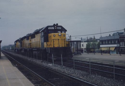 A yellow and black diesel locomotive with the number 5057 and CNW lettering is parked next to a station platform, with buildings in the background under a gray sky.