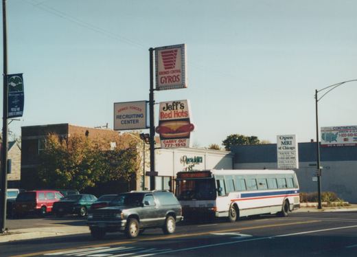 A white Chicago Transit Authority bus with blue and red stripes, fleet number 5412, drives down a street lined with signs for businesses including Kronos-Central Gyros and Jeff's Red Hots. A dark SUV is in the foreground. The sky is clear and the trees have autumn leaves.