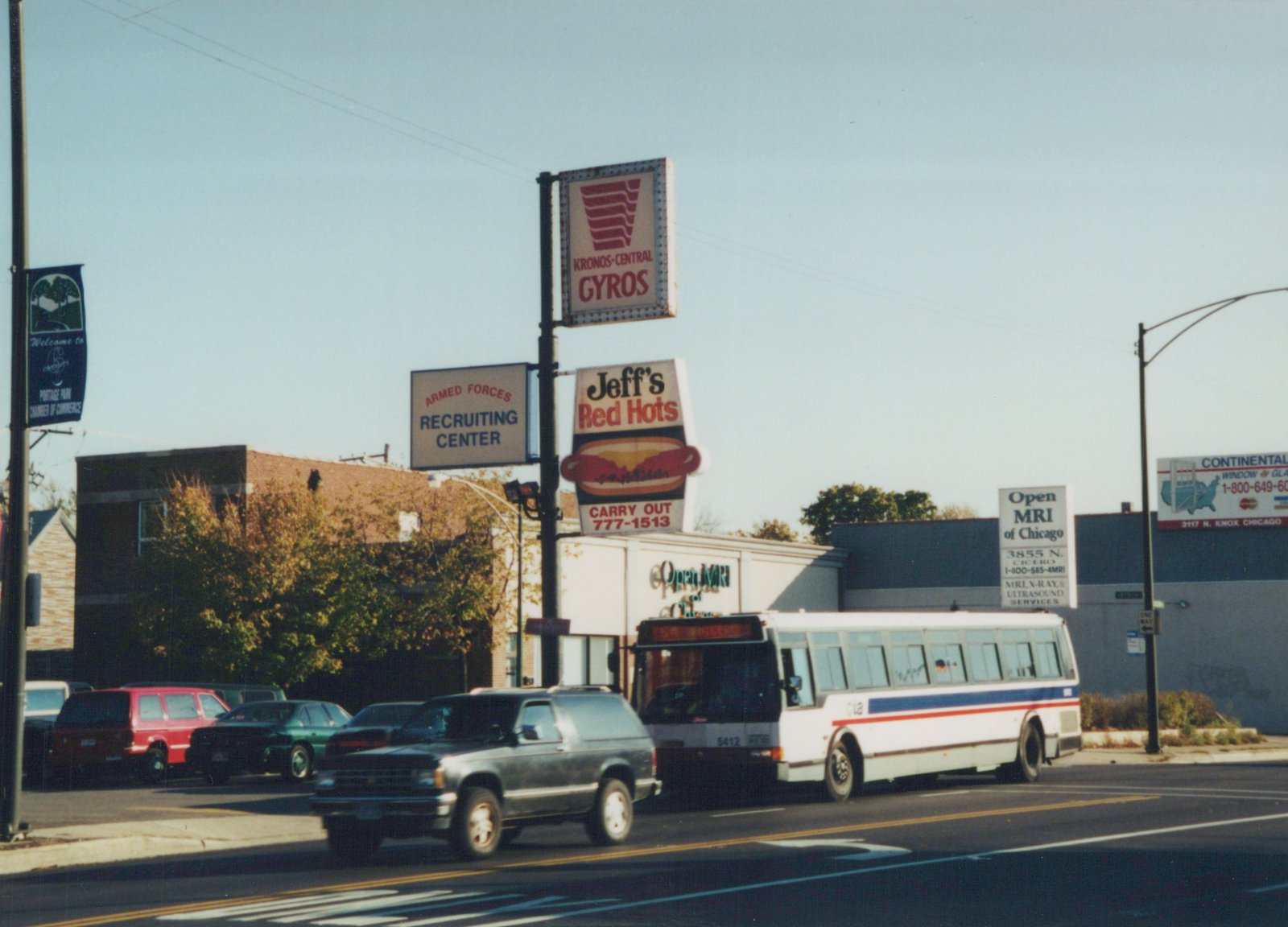 A white Chicago Transit Authority bus with blue and red stripes, fleet number 5412, drives down a street lined with signs for businesses including Kronos-Central Gyros and Jeff's Red Hots. A dark SUV is in the foreground. The sky is clear and the trees have autumn leaves.