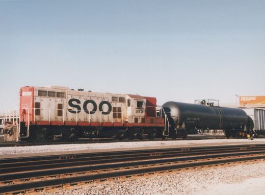 A white and red Soo Line diesel locomotive numbered 383 is coupled to a black tank car on parallel railroad tracks in a yard under a clear blue sky.
