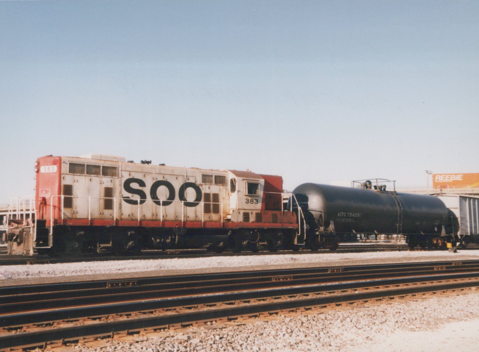 A white and red Soo Line diesel locomotive numbered 383 is coupled to a black tank car on parallel railroad tracks in a yard under a clear blue sky.