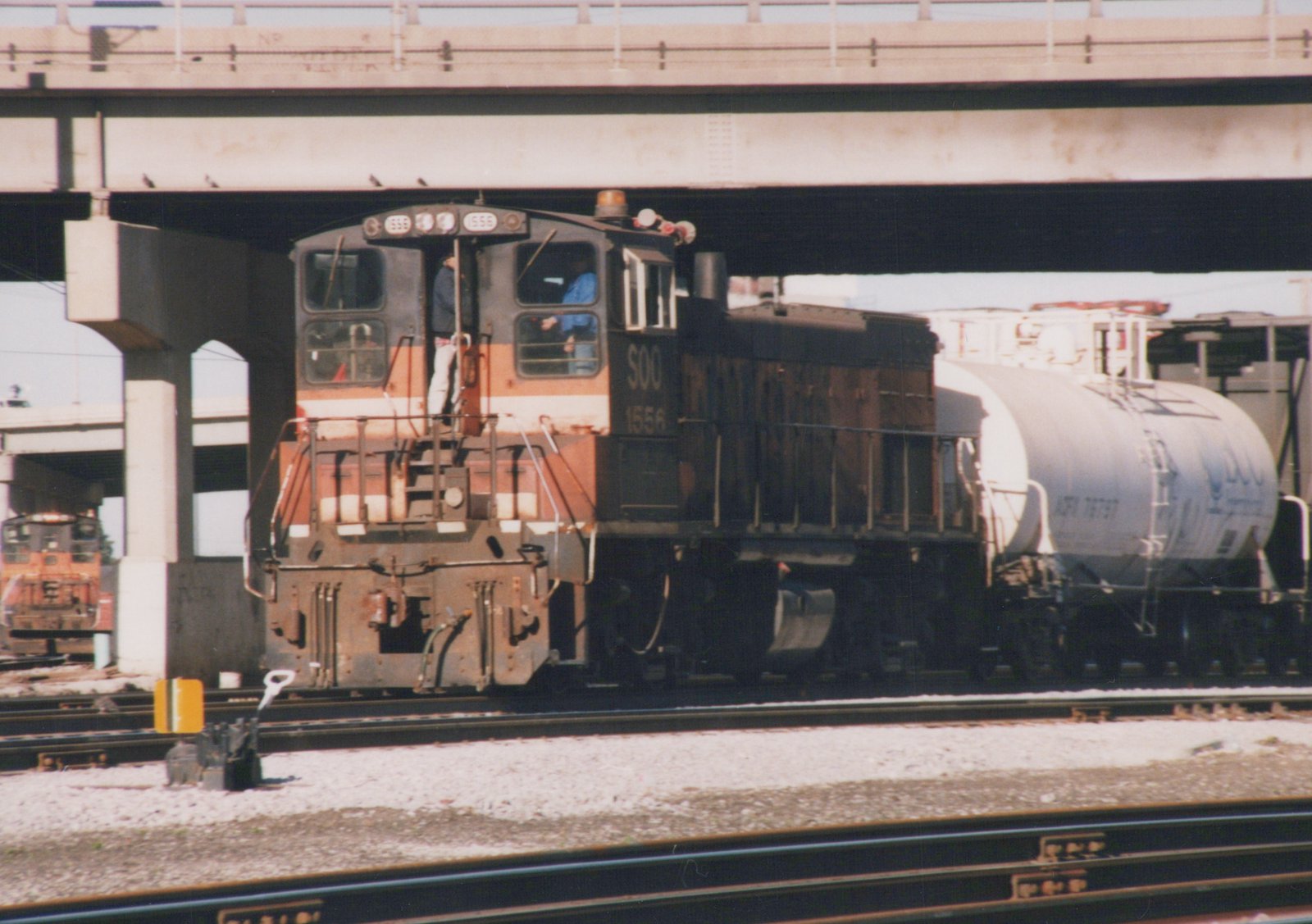 A black, orange and white Soo Line diesel locomotive numbered 1556 is shown in a three-quarter front view, pulling a silver tank car under a bridge in a rail yard on a sunny day.