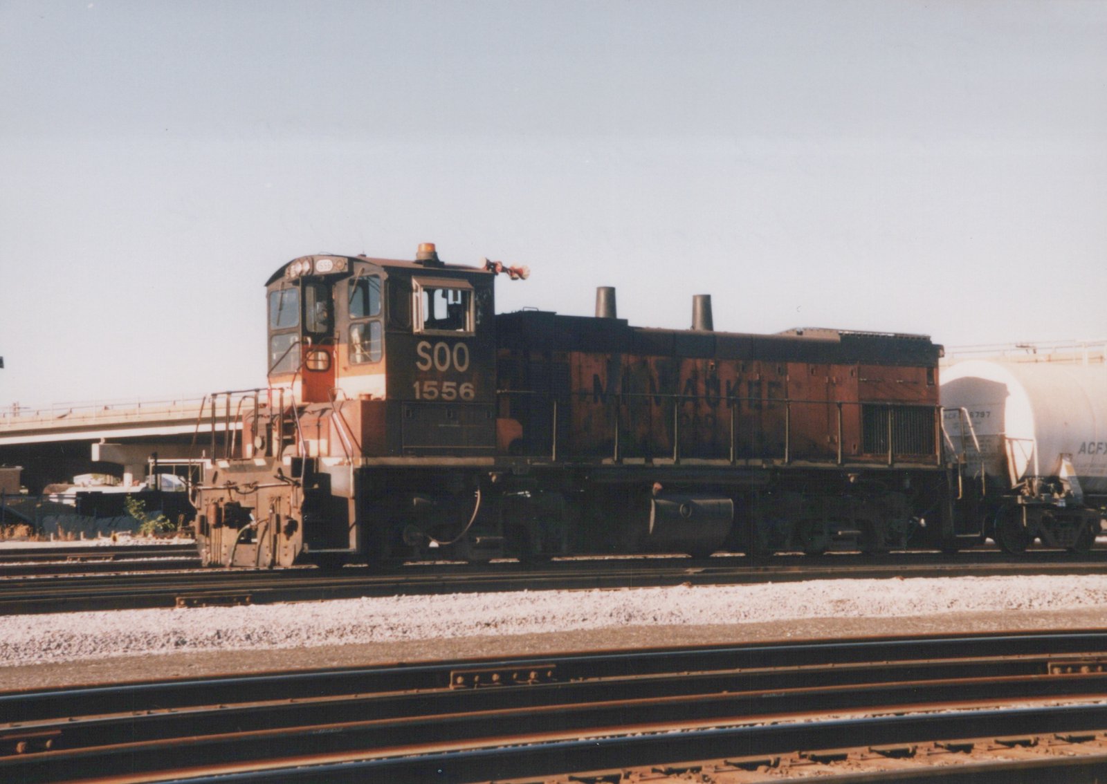 A black and red diesel locomotive with the number 1556 and the letters SOO on the side is stopped on a set of train tracks. Behind it is a white tank car. The setting appears to be a railroad yard with multiple tracks and an overpass in the background.
