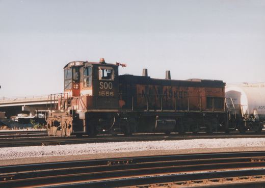 A black and red diesel locomotive with the number 1556 and the letters SOO on the side is stopped on a set of train tracks. Behind it is a white tank car. The setting appears to be a railroad yard with multiple tracks and an overpass in the background.