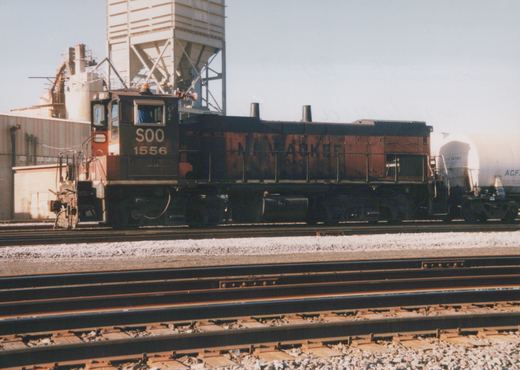 A side view of SOO Line diesel locomotive 1556, showing faded Milwaukee Road lettering on the hood, parked on a railroad track with a white tank car behind and industrial buildings in the background.
