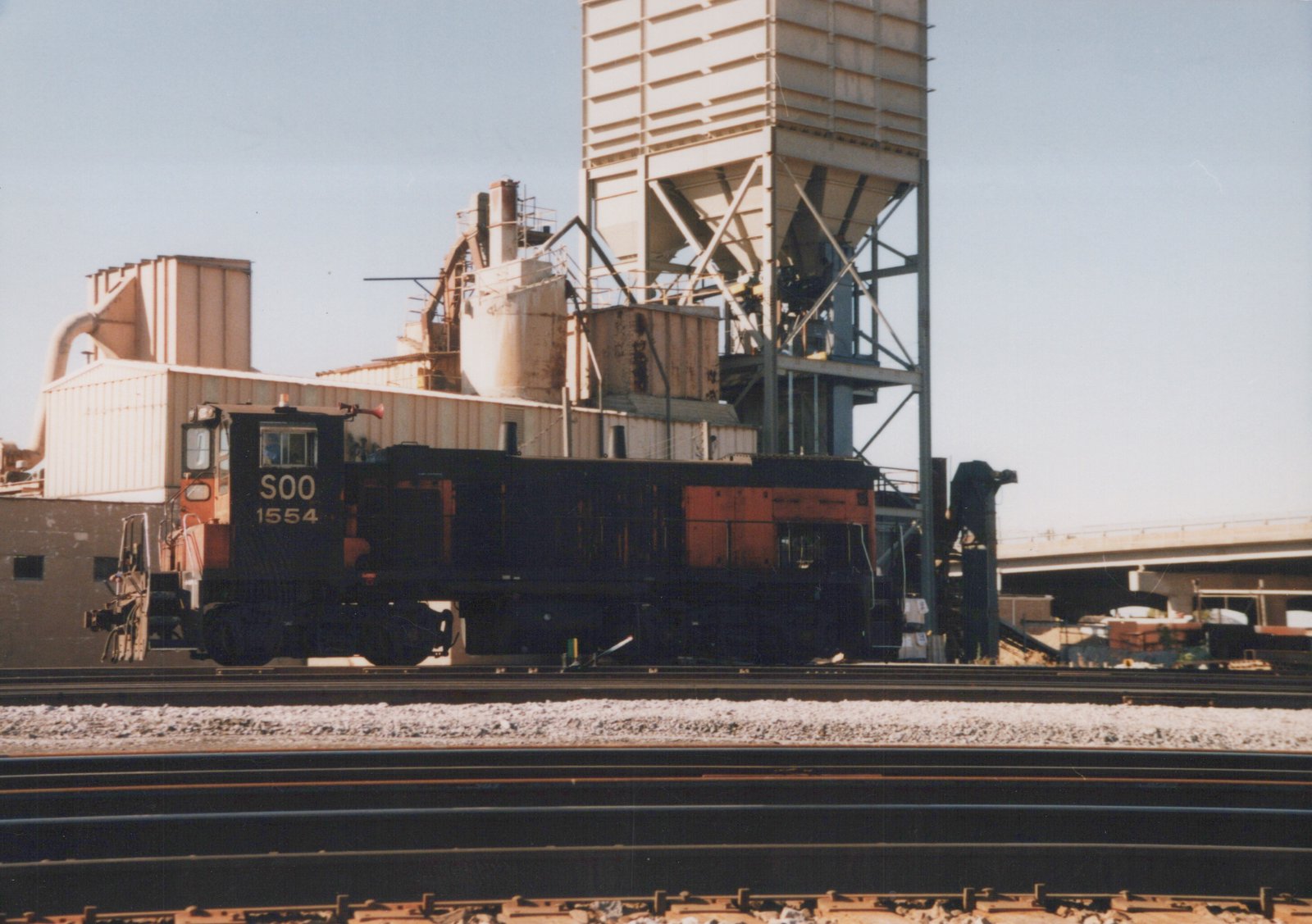 Side view of a black and orange Soo Line EMD SW1200 diesel locomotive numbered 1554, stopped on tracks near industrial buildings and a tall silo structure under a clear sky.