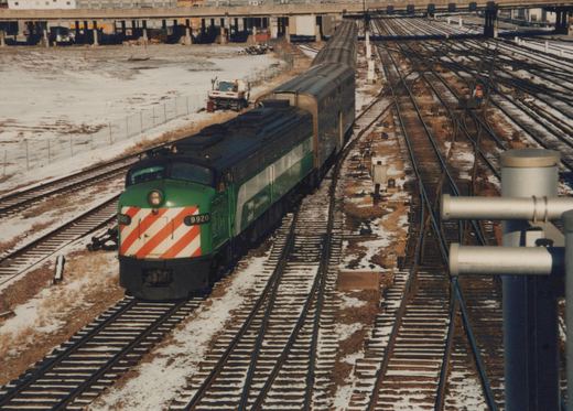 An overhead view of a green Metra train with a red and white striped front, number 9920, traveling on snow-dusted tracks through a complex of multiple railroad lines with a few workers visible on the ground.
