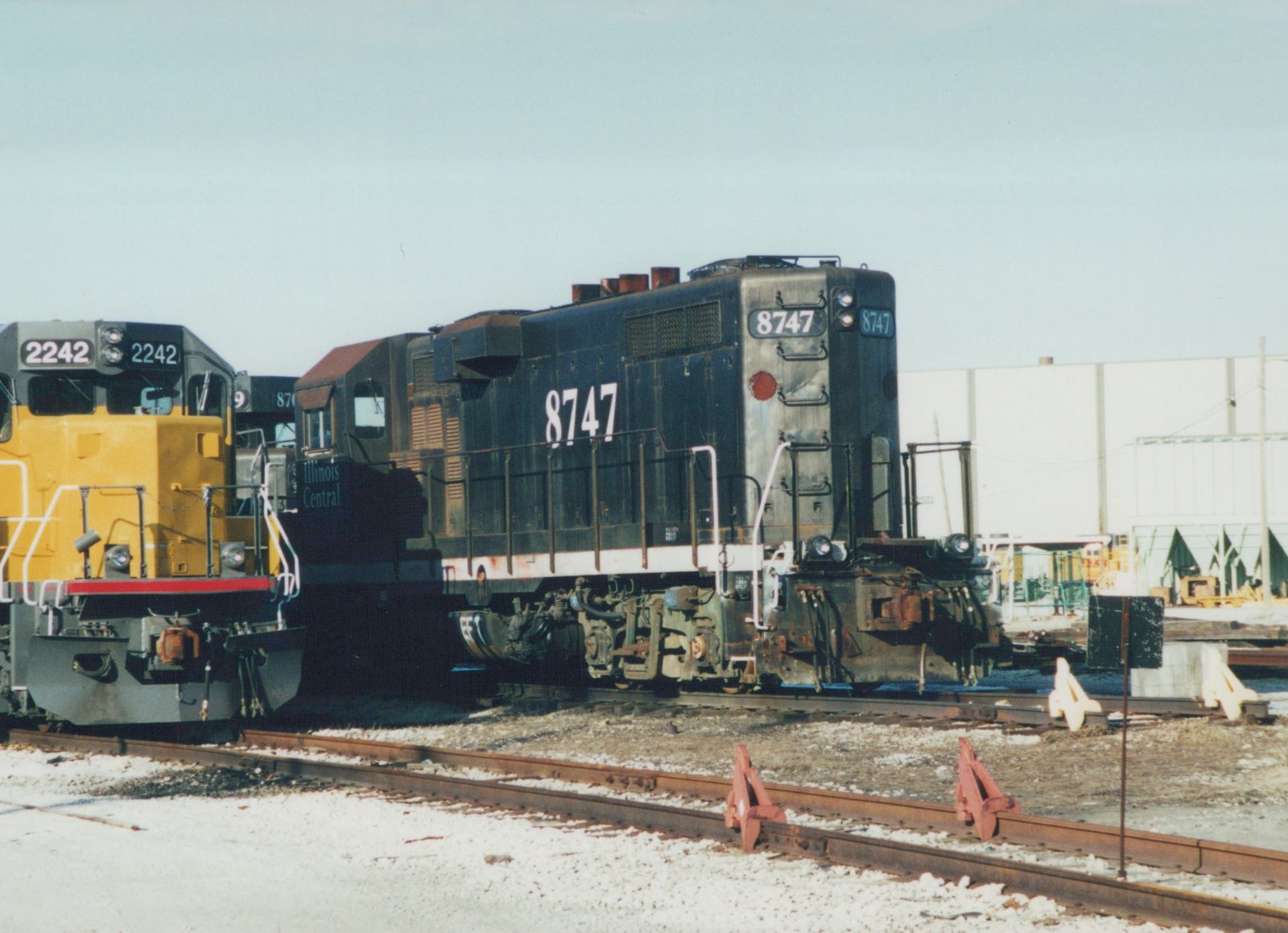 A black and white Illinois Central diesel locomotive, number 8747, is parked on tracks in a snowy yard with a yellow locomotive nearby.